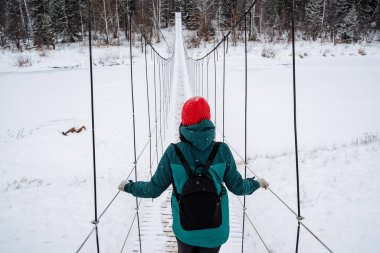 Bir adam donmuş bir nehrin üzerindeki asma köprüde yürüyor. Kışın ormanda daha sık yürüyün, karlı bir ormanda. Çerçeve yukarıda. Yüksek kalite fotoğraf