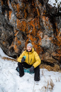 Yakışıklı gezgin çay içer ve uçurumda oturur. Çalı işi. Karlı orman ve kayalar. Piknik ve dağa tırmanma. Vahşi doğada hayatta kalmak. Yüksek kalite fotoğraf