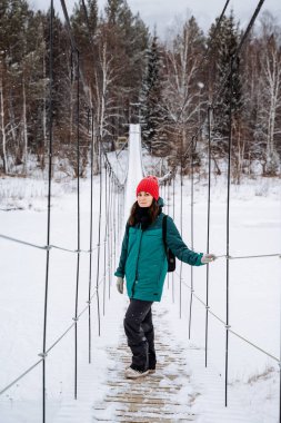 Donmuş bir nehrin üzerindeki asma köprüde duran muhteşem kadın. Kışın ormanda daha sık yürüyün, karlı bir ormanda. Tehlikeli bir halat köprüsünde geçiş. Yüksek kalite fotoğraf