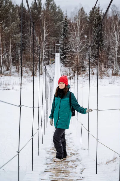Donmuş bir nehrin üzerindeki asma köprüde duran muhteşem kadın. Kışın ormanda daha sık yürüyün, karlı bir ormanda. Tehlikeli bir halat köprüsünde geçiş. Yüksek kalite fotoğraf