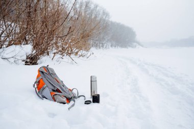 The backpack lies in the snow near the road. The winter trail runs along the river. Equipment for a winter hike. thermos with hot tea. High quality photo