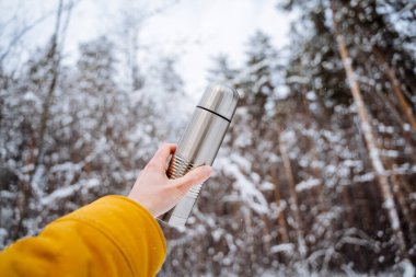 Thermos in hand against the background of the forest. The concept of vacuum objects. Hot tea on a hike in the mountains. Extend your arm up. High quality photo