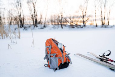 An orange backpack stands on the snow, cross-country skis lie nearby. Countryside scenery. Active lifestyle. High quality photo