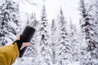 Elinde siyah bir termos var. Metal kapak ve kupa. Kışın yürürken sıcak çay ve kahve içeriz. Sakin bir günde karla kaplı bir orman. Parlak kıyafetler. Yüksek kalite fotoğraf