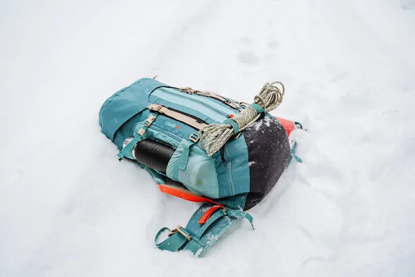 Ένα φωτεινό σακίδιο πεζοπορίας βρίσκεται στο χιόνι. Ένα σχοινί αναρρίχησης είναι δεμένο σε ένα σακίδιο. Στην πλαϊνή τσέπη του θερμός. Πεζοπορία το χειμώνα. Βράχοι και δάση — Φωτογραφία Αρχείου