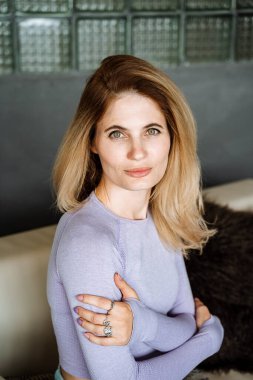 Gri örgülü bluzlu çekici bir sarışın kanepede oturuyor. Kızın tatlı ve çekici gülümsemesi. Parlak yeşil gözler, akılda kalıcı bir bakış. Yüksek kalite fotoğraf