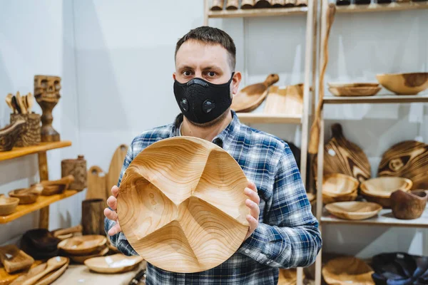 A white man wearing a mask shows off his wares at a craft show. A man holds a round wooden plate in his hands. Carpenter is handmade. High quality photo