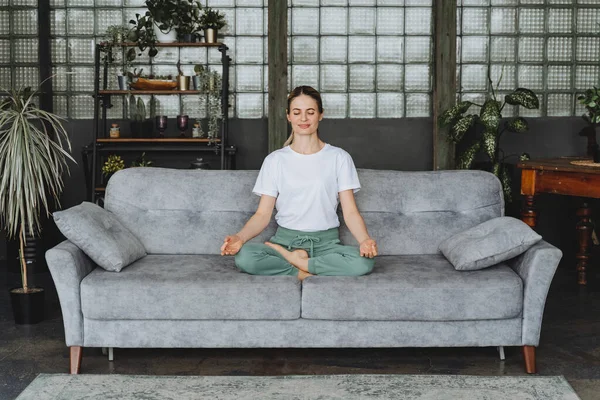 Güzel bir kadın kanepede lotus pozisyonunda oturur. Baddha Konasana. Kendine zaman ayır. Herhangi uygun bir yerde meditasyon. Zen ve sükunet. Yüksek kalite fotoğraf
