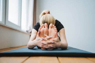 Sarışın bir kız yere uzanır ve bacaklarına uzanır. Yoga yapar ve paspasın üzerindeki tüm kasları gerer. Ayaklara eğilim. PASCHIMOTTANASANA Yüksek kalite fotoğraf