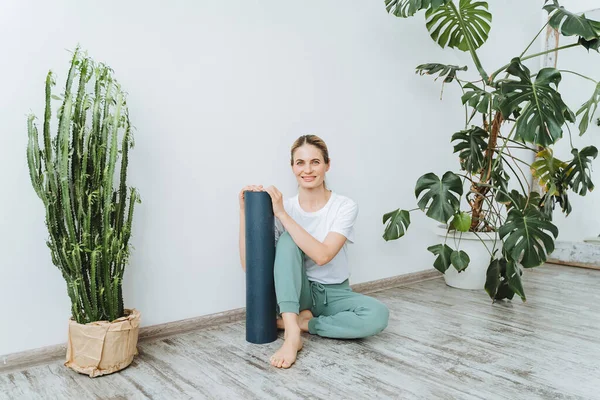 Smiling woman sits on the floor with a rolled up yoga mat. Bright studio for yoga and stretching with plants. Doing yoga for relaxation — Stock Photo, Image