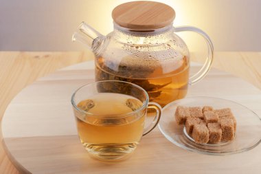 Cam çaydanlıkta sıcak bitkisel çay ve cam bardak, ve tahtadan bir tabakta kahverengi şeker parçaları..