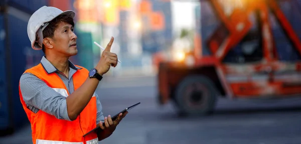 Asian male industrial engineer in white helmet, orange safety vest holding tablet, He\'s checking the number of containers for accuracy. Large warehouse management specialists in container terminal.