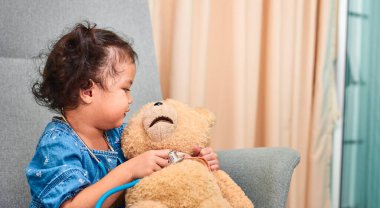 Cute curly haired Asian girl, about 3 years old with stethoscope plays with teddy bear while sitting on the chair in the living room.