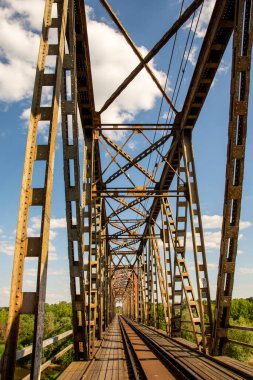 The metal structure of the railway viaduct over the river against the background of a blue sky with clouds. Day. 