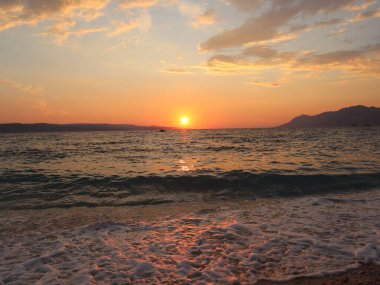 Deniz kıyısında gün batımı, batan güneş ıslak kuma ve suyun yüzeyine yansıyor. Gün batımında deniz manzarası, gökyüzü, deniz ve deniz köpüğü batan güneşin turuncu ışınlarıyla aydınlatılır.