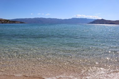 Hırvatistan 'ın Baska kentinde, güneşli bir günde Krk adasında bir plaj. Açık turkuaz suyu ve ufukta bir dağ silsilesi olan bir tatil köyündeki resim gibi bir sahil.