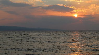 Deniz üzerinde pembe bronz tonlarda gün batımı, Brela, Hırvatistan. Turuncu güneş bulutların ardında batar, güneş ışınları bir yaz akşamı denizin yüzeyine yansımaktadır.