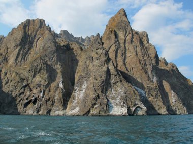Kayalar ve deniz, sönmüş olan Karadağ volkanının dağ yamaçları beyaz bulutlar ve mavi deniz ile parlak mavi bir gökyüzünün arka planına karşı. Deniz manzarası, Karadağ dağlarının yamaçları bulutlu ve güneşli bir yaz gününde deniz manzarası, Kırım.