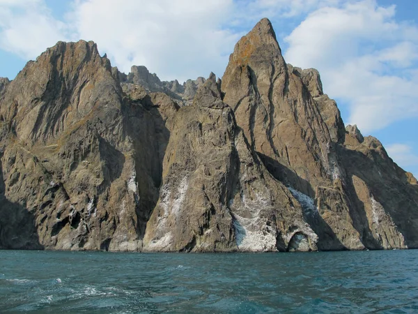 Kayalar ve deniz, sönmüş olan Karadağ volkanının dağ yamaçları beyaz bulutlar ve mavi deniz ile parlak mavi bir gökyüzünün arka planına karşı. Deniz manzarası, Karadağ dağlarının yamaçları bulutlu ve güneşli bir yaz gününde deniz manzarası, Kırım.