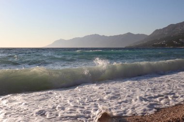 Bir yaz günü fırtına sırasında sahil ve deniz. Denizde dalgalar, deniz kıyısında güneş ışınlarında deniz köpüğü, Hırvatistan 'da plaj tatili.