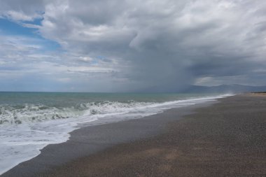 Ufukta deniz üzerinde yağmur, bulutlu gökyüzü ve yağmurlu bir günde sahilde dalgalar. Manzaralı deniz manzarası yağmur bulutlarının arka planında, mavi gökyüzü Calabria, İtalya sahillerinde bir çakıl taşı kumsalında