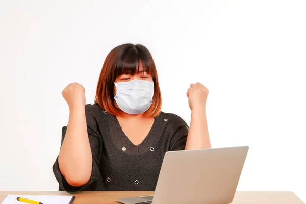 Concept work at home : Fat Asian woman wearing a coronavirus protective mask (covid-19), sitting on a desk, using a laptop computer, expressing gratitude with her work to achieve her goals.