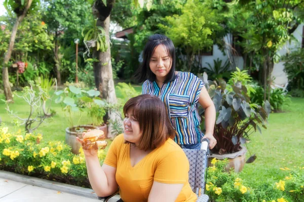 Fat woman sitting in a wheelchair, She likes to eat pork hamburgers, ignoring the girl friend who looks with disgust at not refusing to stop eating.