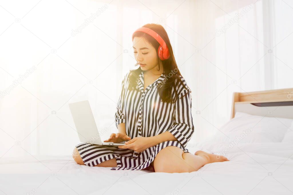 Casual work at home concept : Asian teenage woman wearing pajamas on the bed, woke up early to work, wearing headphones, sounds from a laptop computer in the morning of the weekend in her bedroom.