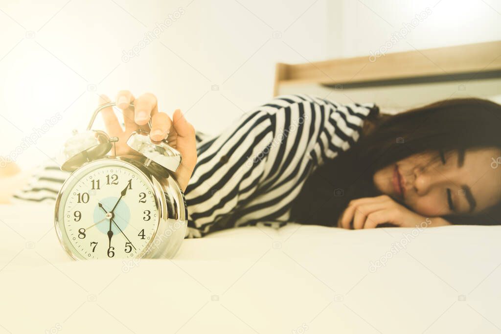 Young woman lying in bed reaches out to turn off the sound of the alarm clock at 6.00 AM in the morning ; Sleepy girl lying in bed reaching to turn off the alarm of the clock.