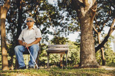 Görme engelli yaşlı ve yaşlı Asyalı bir adam parktaki tahta bir sandalyede oturuyor. Duyularını rahatlatmak ve doğanın seslerini dinlemek için..