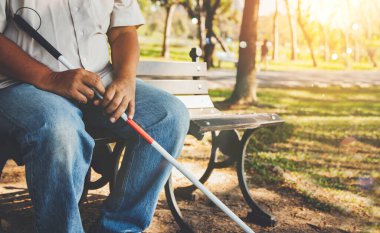 Elderly blind man with a blind cane sits alone on a park bench : Lind man who wants peace and lives in solitude concept. clipart