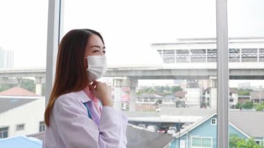 Üniformalı güzel kadın doktor, Coronavirus, COVID-19 ve yeni nesil Omicron 'un hızla yayılmasını önlemek için hastane ofisinde bir pencerenin önünde tıbbi maske takıyor..