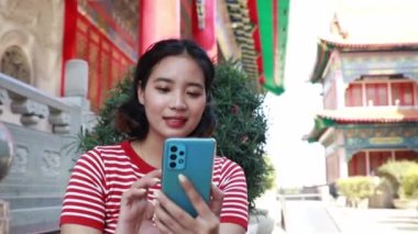 Kırmızı tişörtlü Asyalı bir kadın, Çin Yeni Yılı 'nda bir tapınağı ziyaret eder, bir sandalyeye oturur, akıllı telefonuyla oynar ve neşeli, neşeli bir yüzle kadrana bakar..