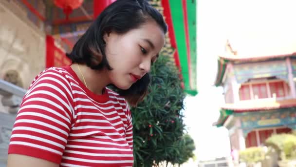 身穿鲜红色衣服的年轻亚洲女人坐在智能手机上 然后看着相机 面带微笑 心地善良 — 图库视频影像