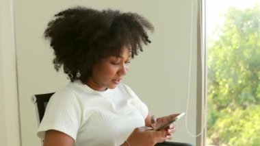 Afrika kökenli Amerikalı bir kadın oturma odasında akıllı telefonuna oturmuş komik bir yüzle kameraya gülümsüyor..