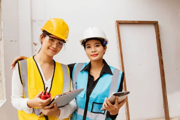 Portrait two beautiful asian engineers young women architects and workers work together wearing helmets and life jackets holding laptops and tech equipment on the construction site : Teamwork concept.