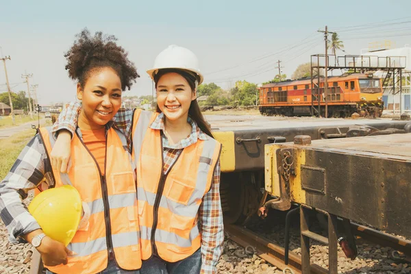 Portrait two engineer industrial worker two young women from different nationalities and cultures beaming shoulders at the camera looking happy friendship featured at work diesel rail train station.