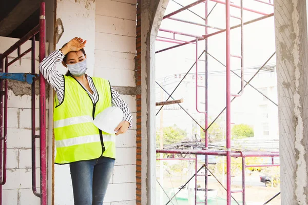 Asian female architect engineer wearing protective mask working tired and worried about the construction site from the COVID-19 outbreak. This caused the workers to infect and delay the construction.