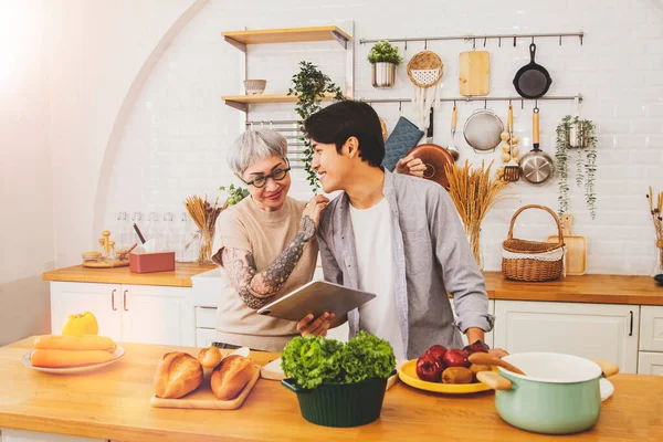 Couple between an old woman tattooed on the arm and handsome childhood boy who bond and love each other well, embrace close care, cook in the kitchen with joy : Age difference and love concept.