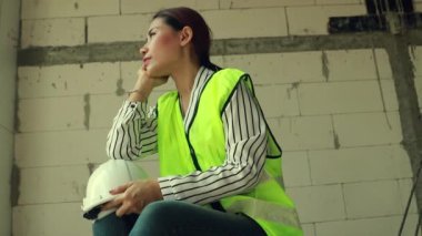 Güzel Asyalı kadın ustabaşı mimar mimar miğfer kırma şantiyesinde oturuyor sakince bir şeyler düşünüyor, kameraya bakıyor..