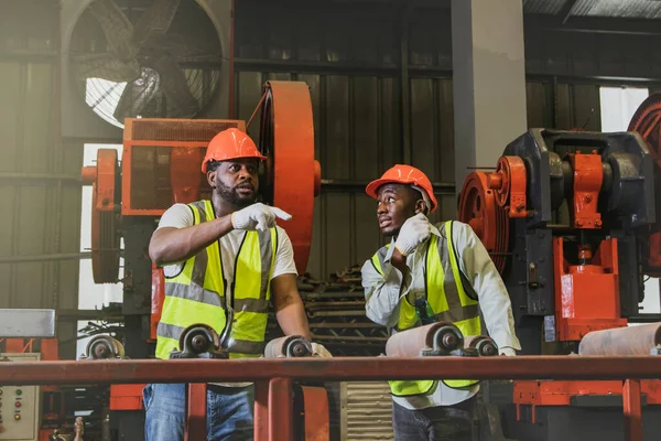 Two African American workers in helmets stand together discussing production explaining sharing brainstorming sessions at the metal sheet manufacturing plant to make it work smoothly and safely.