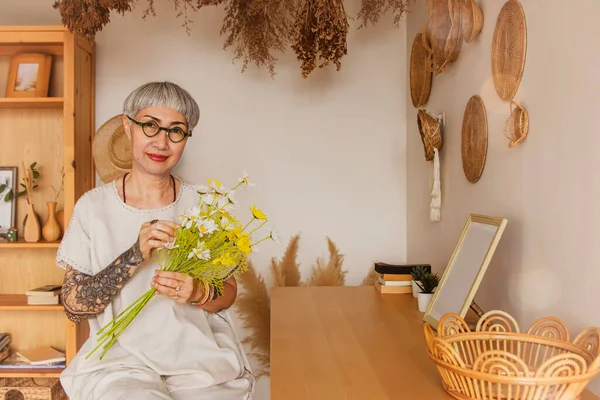 Beautiful retired 60 year old asian woman with short gray hair wearing glasses healthy dazzling clear face charming relaxed lively arranged flowers at home smiling happily looking at the camera.