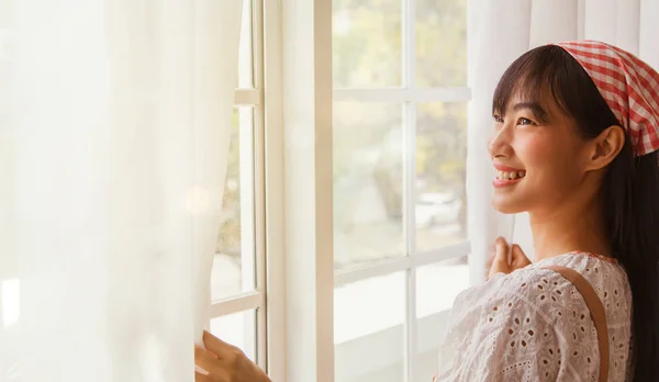 Good morning new day : Asian cute girl fairy opens the window curtain greets the morning sun in the living room looks outside smiling has a good attitude and health.