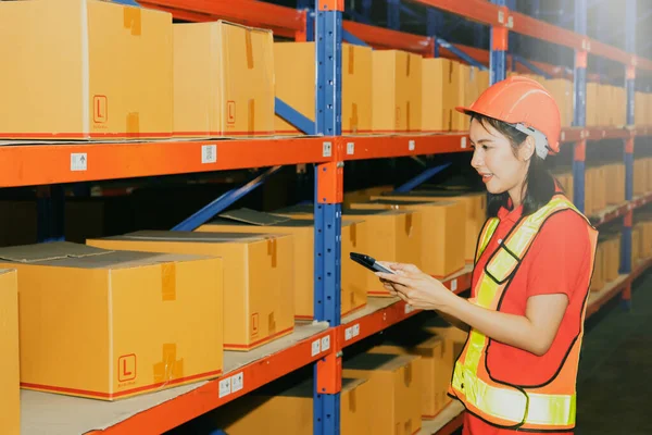 Side view beautiful young asian female worker working the night shift wearing a helmet works with smiling face, use tablet to inspect the boxes from the warehouse shelf to verify they are correct.