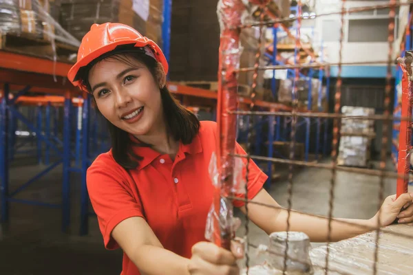 Side view beautiful young asian female worker working the night shift wearing a helmet works with smiling face, use tablet to inspect the boxes from the warehouse shelf to verify they are correct.