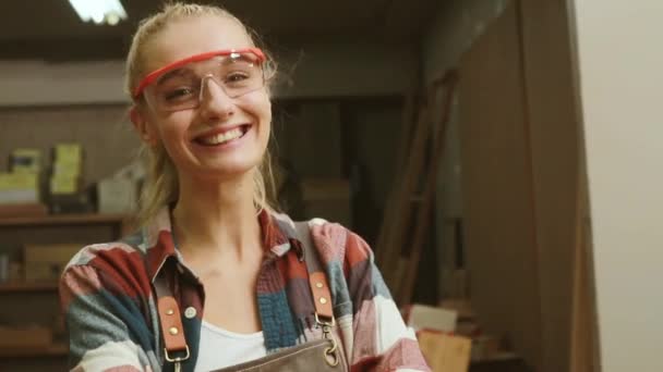 一家家具厂的女木匠戴着安全玻璃 自豪地站在一个木工车间里 手里拿着一个看上去很聪明的相机 双手交叉 竖起大拇指 — 图库视频影像