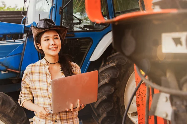 Asian woman farmer smile, agribusiness and livestock operator holding laptops to inspect machines and tractors for agricultural use, inspect maintenance services and use on agricultural plantations.