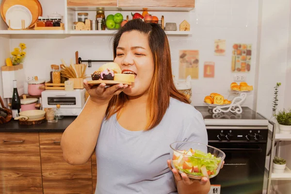 Blissful fat asian woman prefers sugary and buttery bakery desserts by holding bowl of healthy salad, but she doesn\'t : Unhealthy nutrition concept