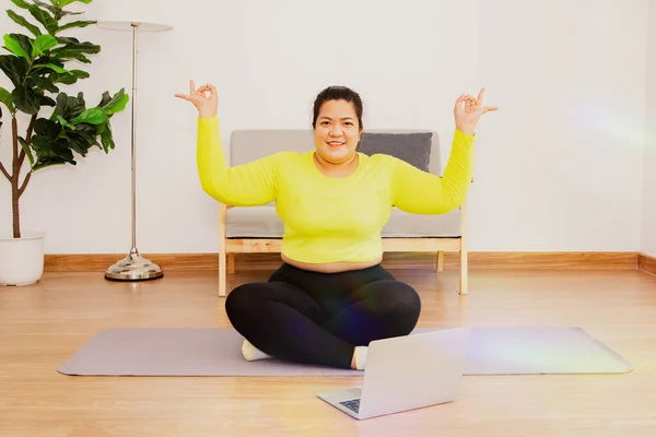Fat woman exercising online to lose weight sitting meditation yoga poses calm and healthy relax take time to take care of well being wearing sportswear yellow shirts spot yoga at home.
