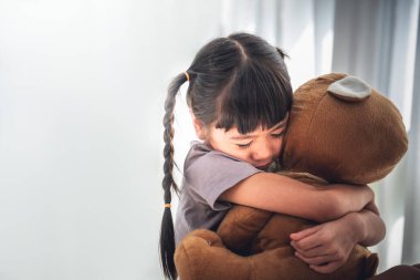 Asyalı kızın bulanık görüntüsü 6 yaşında, büyük kahverengi bir ayıya sarılıyor, yalnızlık ve üzüntü içinde, beyaz sırtında, çocuklara ve aile kavramına.